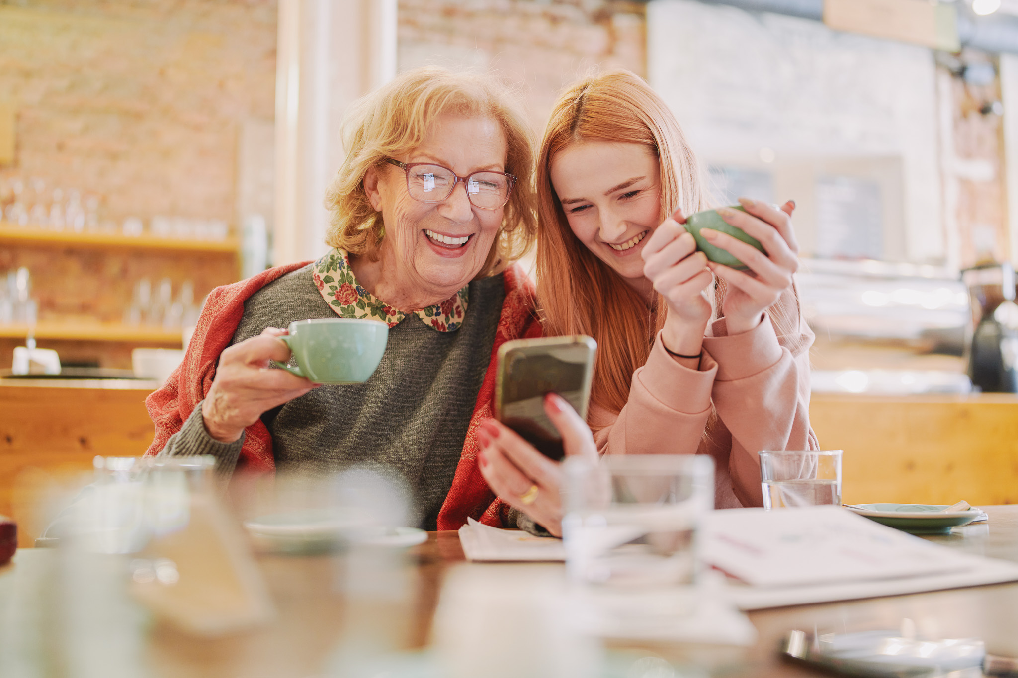 Ältere Frau sitzt mit jungem Mädchen am Tisch mit Kaffeetassen und einem Smartphone